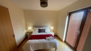 Une chambre à coucher avec un lit, deux tables de chevet avec des lampes, un plaid rouge et une grande fenêtre avec des stores.