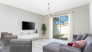 A modern living room with a grey sofa, a flat-screen TV mounted on the wall, a dining area with chairs, and a large sliding glass door leading to a balcony.