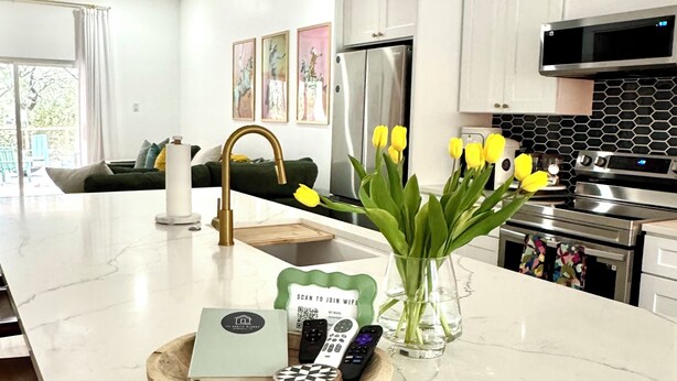 A modern kitchen with white cabinets, a stainless steel refrigerator, and an oven. There is a marble countertop with a tray holding a remote control, a book, and a vase with yellow flowers. - Charming 3-bedroom farmhouse in Wimberley with AC, WiFi (Wimberley)