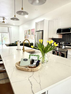 A modern kitchen with white cabinets, a stainless steel refrigerator, and an oven. There is a marble countertop with a tray holding a remote control, a book, and a vase with yellow flowers. - Charming 3-bedroom farmhouse in Wimberley with AC, WiFi (Wimberley)