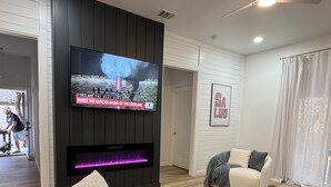 A modern living room with a large flat-screen TV mounted on a black panel wall, a fireplace below it, a green sofa, a white armchair, and a ceiling fan. - Charming 3-bedroom farmhouse in Wimberley with AC, WiFi (Wimberley)
