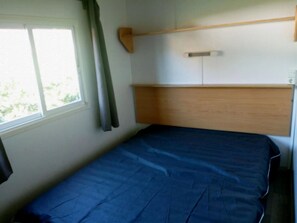 Una habitación pequeña con una cama individual, cabecera de madera, ventana con cortinas y estante fijado a la pared.