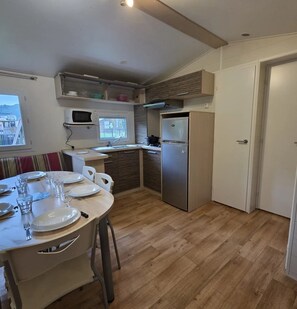 A compact kitchen with a dining area, a microwave, a refrigerator, and a window overlooking a balcony. - ZEcamping 4* animé piscine couverte chiens acceptés locatif TV 32m2 (Vic-sur-Cère)