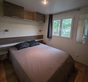 A bedroom with a bed, a window with curtains, a bedside table, and a wall-mounted shelf. - ZEcamping 4* animé piscine couverte chiens acceptés locatif TV 32m2 (Vic-sur-Cère)