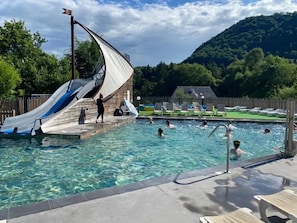 A swimming pool with a slide, surrounded by lounge chairs and a wooden deck. - ZEcamping 4* familial piscine couverte pet friendly mobil-home 22m2 (Vic-sur-Cère)