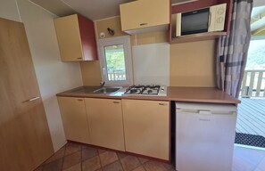 A compact kitchen with a sink, stove, and microwave, featuring a window and a view of the outdoors. - ZEcamping 4* familial piscine couverte pet friendly mobil-home 22m2 (Vic-sur-Cère)