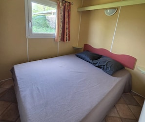A bedroom with a bed, a window with curtains, a bedside table, and a wall-mounted light fixture. - ZEcamping 4* familial piscine couverte pet friendly mobil-home 22m2 (Vic-sur-Cère)