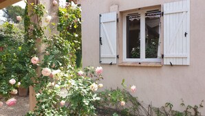 Un bâtiment doté d’une fenêtre ornée de volets blancs, entouré d’un jardin où s’épanouissent des rosiers roses.