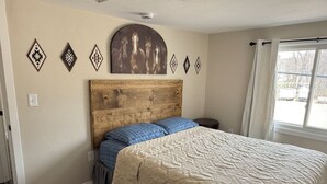 Une chambre à coucher avec une tête de lit en bois, un lit avec une literie bleue et beige, une fenêtre avec des rideaux et des œuvres d’art décoratives au mur.