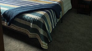 A bedroom with a wooden ceiling, a bed with a striped blanket, two pillows, a nightstand with a lamp, and a painting of a sunset over a pier. - Bootjack Lake House: Private Lakefront in the Heart of the Keweenaw (Lake Linden)