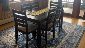 A dining area with a wooden table and chairs, a patterned rug, and large windows with a view of a body of water. - Bootjack Lake House: Private Lakefront in the Heart of the Keweenaw (Lake Linden)
