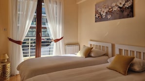 Habitación de hotel con dos camas, una ventana con cortinas traslúcidas, un radiador, un cuadro colgado en la pared y una mesita de noche con una lámpara.