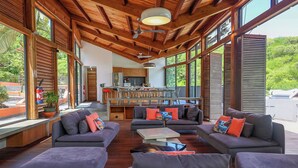 Un vaste espace de vie agrémenté de canapés et de poufs gris, d’un plafond en bois et de grandes fenêtres offrant une vue sur la verdure.