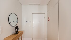 A hallway with a wooden floor, a round mirror on a wooden console, a white door with a handle, and a fire alarm on the wall.