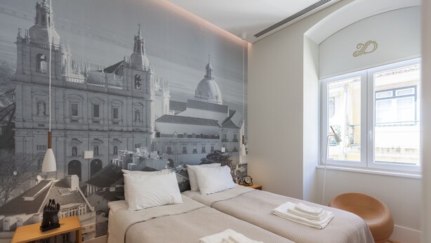 A hotel room with two beds, a mural of a cityscape, a window with a view, and a small wooden side table.