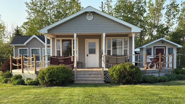 A two-story house with a front porch, a small deck, and a fenced yard. - Cozy, family and pet friendly cottage that is steps to a beautiful PEI beach. (Bonshaw)