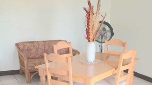 Un'area pranzo con un tavolo e sedie in legno, un vaso con piante essiccate, una ventola a soffitto e un divano decorato sullo sfondo.