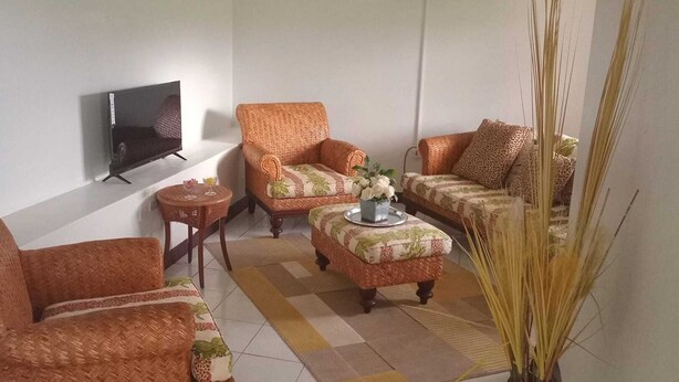 Un soggiorno con una TV a schermo piatto montata a parete, due poltrone in vimini, un divano decorato con motivi, un tavolino tondo con un bicchiere e un vaso ornamentale con fiori.