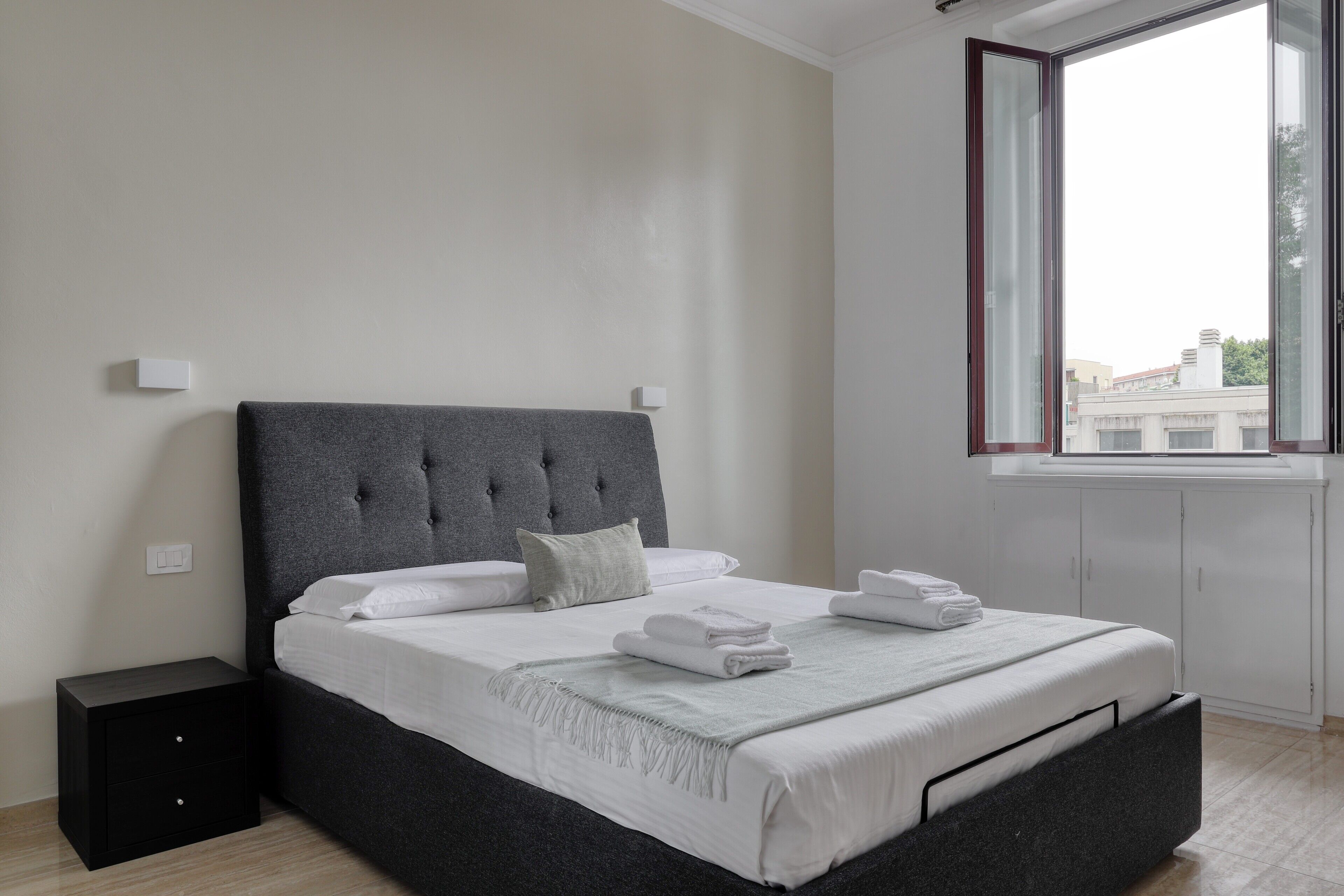 A modern bedroom with a large bed, a nightstand, a window with a view of buildings, and a white wall.