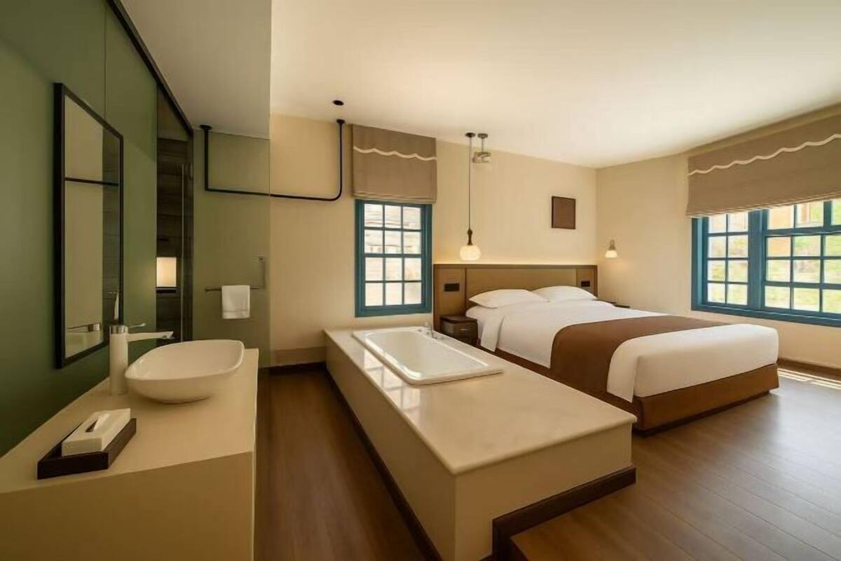 A modern hotel room with a large bed, a separate bathroom with a sink and mirror, and large windows.