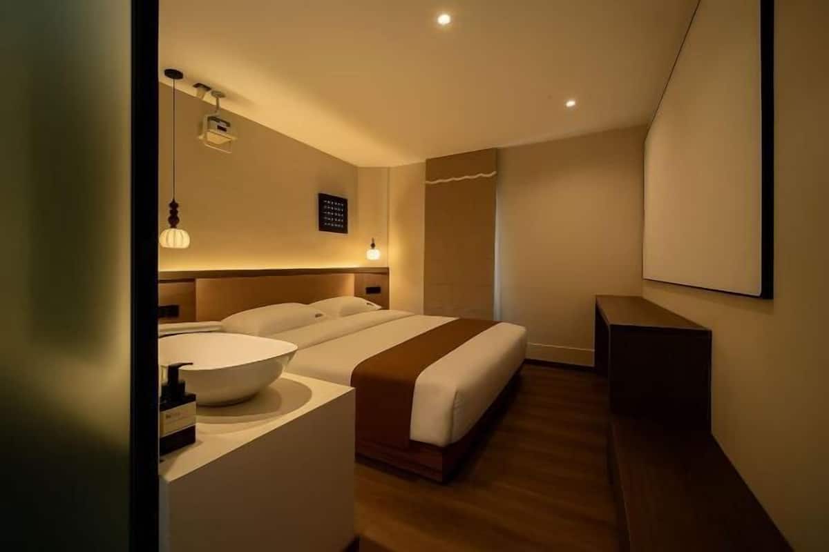 A modern hotel room with a large bed, a bedside table with a bowl sink, and a small desk.