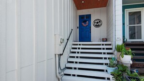Uma escadaria leva a uma porta azul decorada com uma guirlanda, paredes brancas e plantas em vasos.