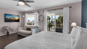 Quarto com cama grande, ventilador de teto, televisão, varanda com cortinas e planta em vaso.