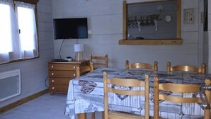 Una habitación con techo de madera, televisor montado en la pared, mesa de comedor con sillas, cómoda y ventana con cortinas.