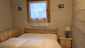 Un dormitorio con cama de madera, una mesita de noche con lámpara, una ventana con cortina y dos pequeños cuadros enmarcados en la pared.