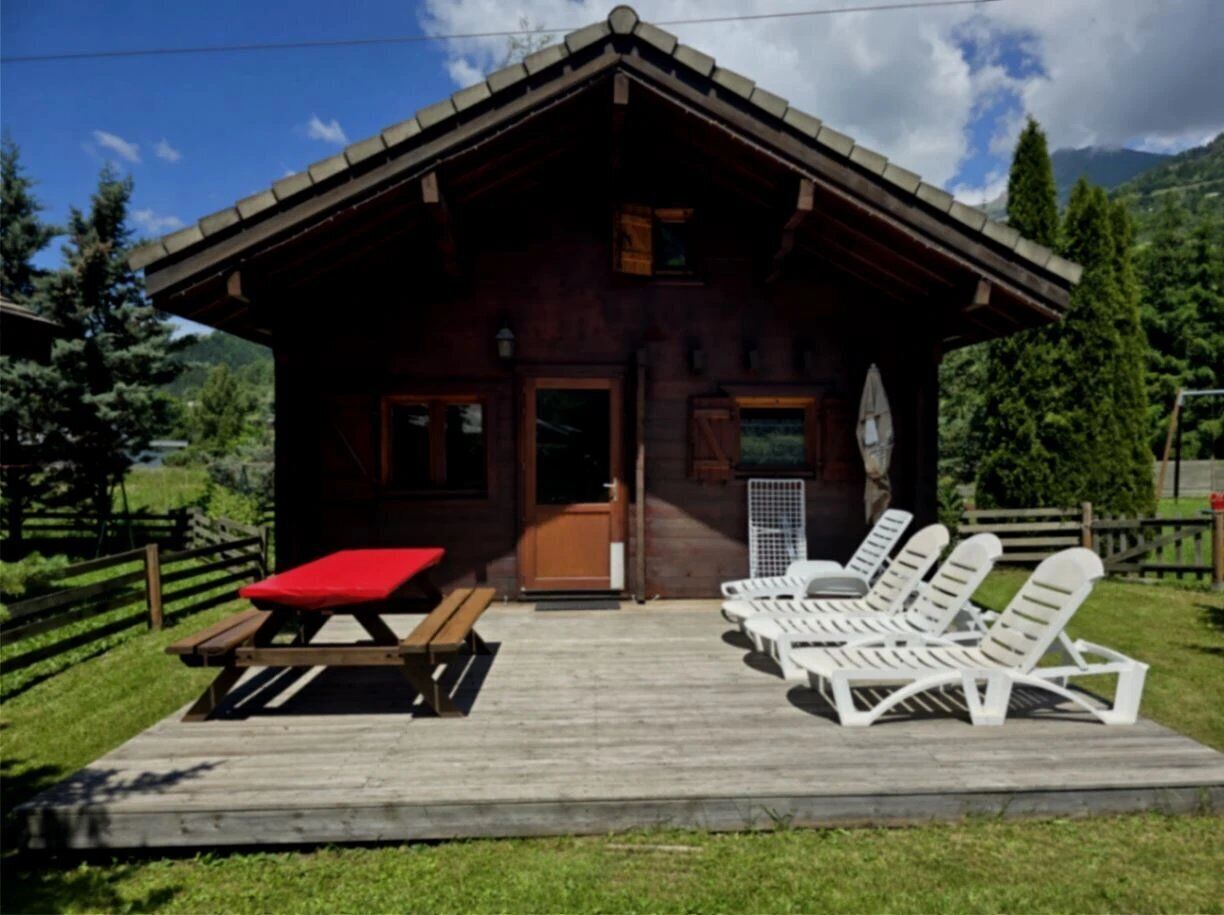 Une cabane en bois avec une véranda, une table de pique-nique et des chaises longues blanches.