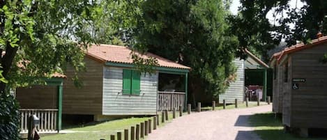 A row of wooden cabins with green shutters, a gravel path leading to them, and a sign indicating a disabled accessible toilet.