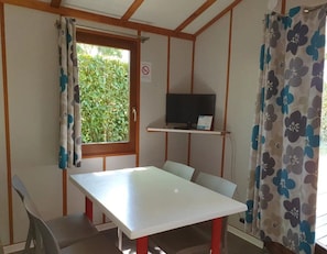 Quarto com uma mesa branca, televisão em uma estante, uma cadeira e uma janela com cortinas.
