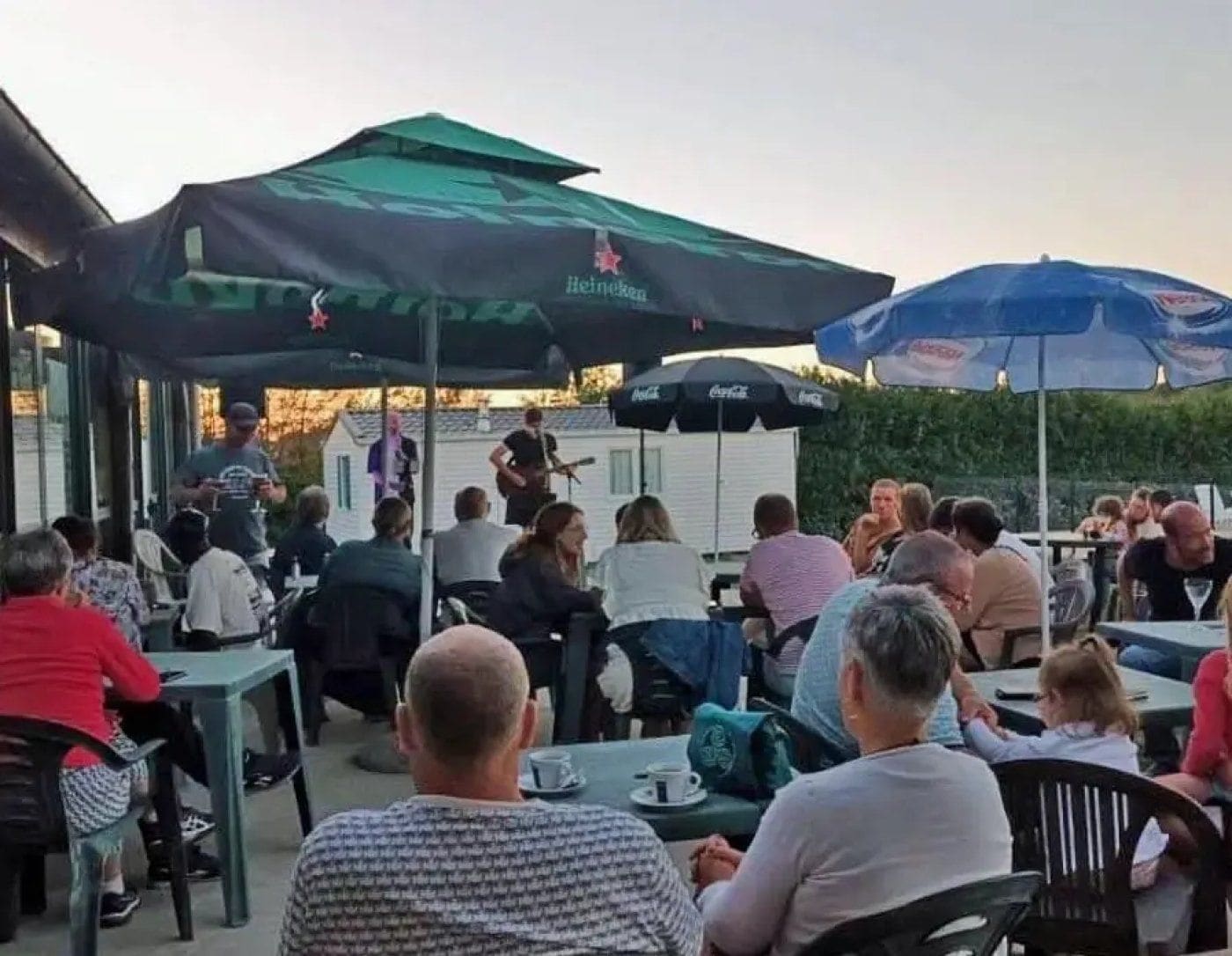 An outdoor seating area with tables and chairs, covered by umbrellas, and a stage with a band performing.
