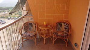 A balcony with two wicker chairs and a small round table, overlooking a dirt road with vehicles.