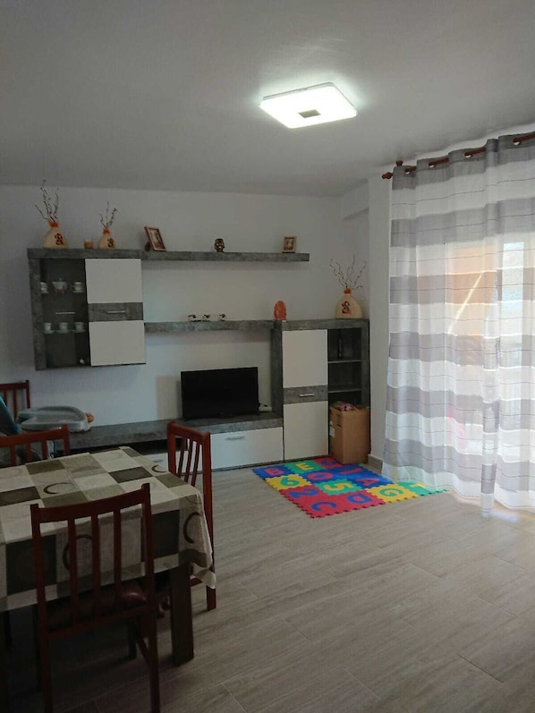 A living room with a TV, a dining table with chairs, a colorful play mat, and a window with curtains.