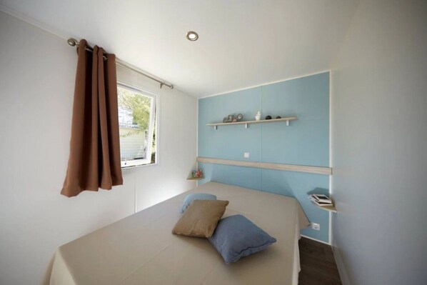A bedroom with a bed, a window with curtains, a shelf with decorative items, and a wall-mounted light fixture. - ZEcamping 3* piscine couverte dog friendly animations locatif TV 27m2 (Avrillé)