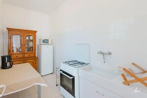 Uma cozinha compacta com fogão a gás, micro-ondas e frigorífico. Há um armário de madeira com portas de vidro e uma pequena mesa com uma chaleira.