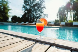A glass of orange juice with a slice of orange on a wooden deck by a pool.
