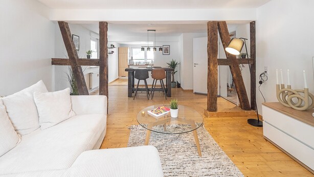 Ein modernes Wohnzimmer mit einem weißen Sofa, einem Glastisch und Holzboden. Im Hintergrund ist ein Essbereich mit Tisch und Stühlen zu sehen.