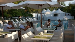 Ein Essbereich am Pool mit weißen Tischen und Stühlen, Sonnenschirmen und einem Pool im Hintergrund.