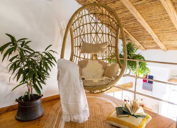 Ein Zimmer mit einem Hängekorbsessel, einem Blumentopf, einem Teppich und einem Bücherregal mit Büchern und einem kleinen Tisch.