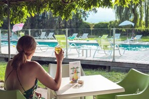 A woman sitting at a table by a poolside, holding a glass of beer, with a menu and a small bowl of snacks on the table.