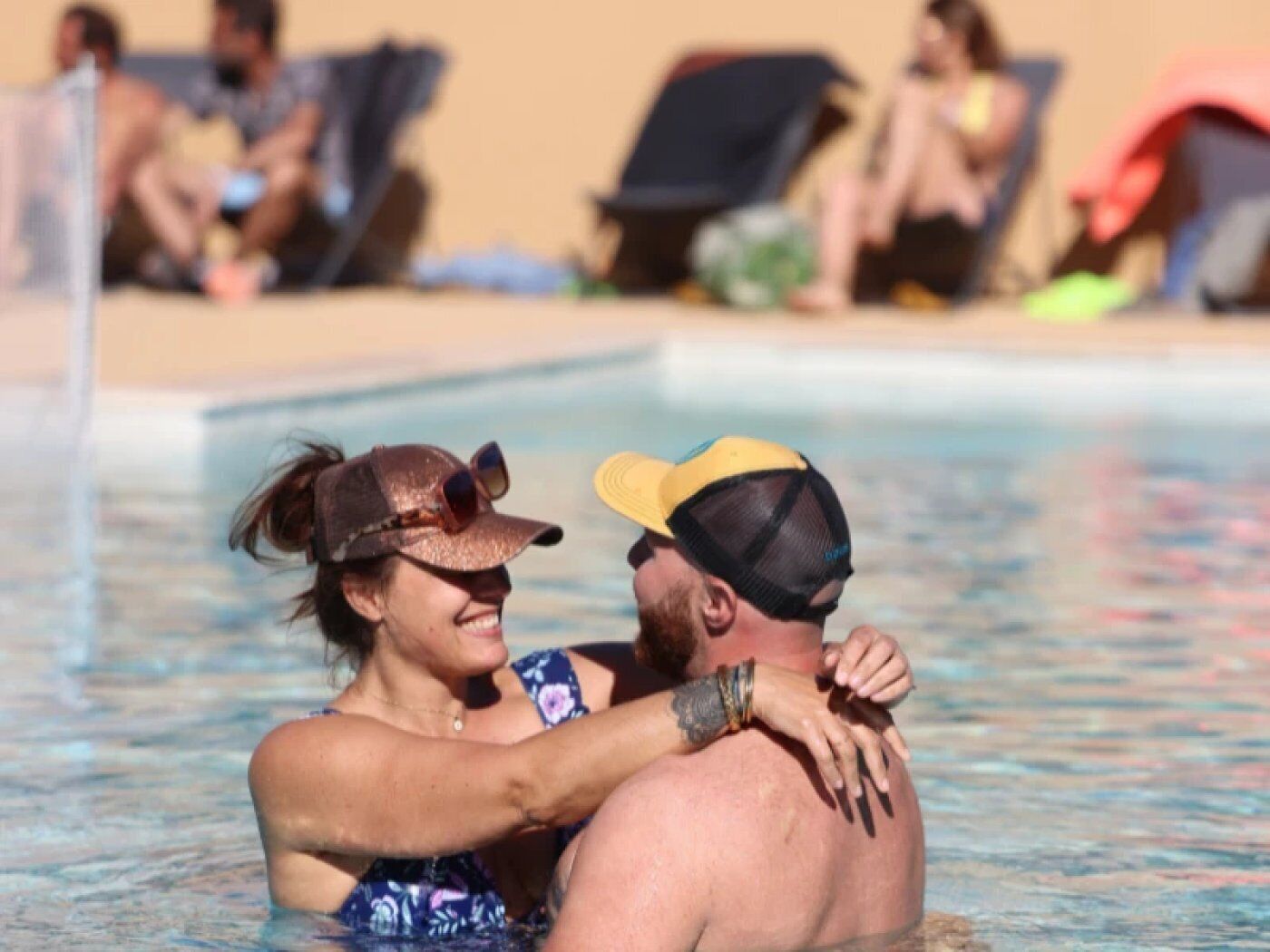 A couple embracing in a swimming pool with other people in the background.