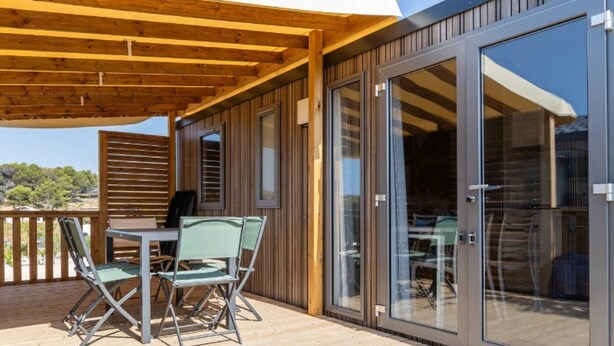 A wooden deck with a table and chairs, a glass door, and a view of the surrounding landscape. - ZEcamping 3* piscine chauffée locatif TV LV BBQ 33m2 6 pers (Martigues)