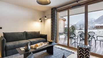 Un salon moderne avec un canapé gris, une table basse sur laquelle se trouve un plateau avec du pain et une tasse, et une vue sur un espace extérieur enneigé aménagé avec des tables et des chaises.