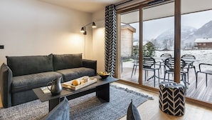 Een moderne woonkamer met een grijze bank, een bijzettafeltje met een plank brood en een kopje, en uitzicht op een besneeuwde buitenruimte met tafels en stoelen.