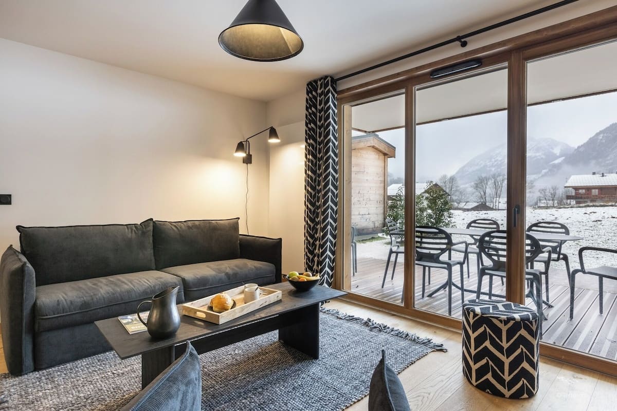 A modern living room with a grey sofa, a coffee table with a tray of bread and a cup, and a view of a snowy outdoor area with tables and chairs.
