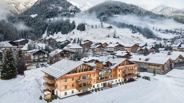 Een besneeuwd bergdorp met houten gebouwen, waaronder een groot hotel met meerdere balkons en een piste op de achtergrond.