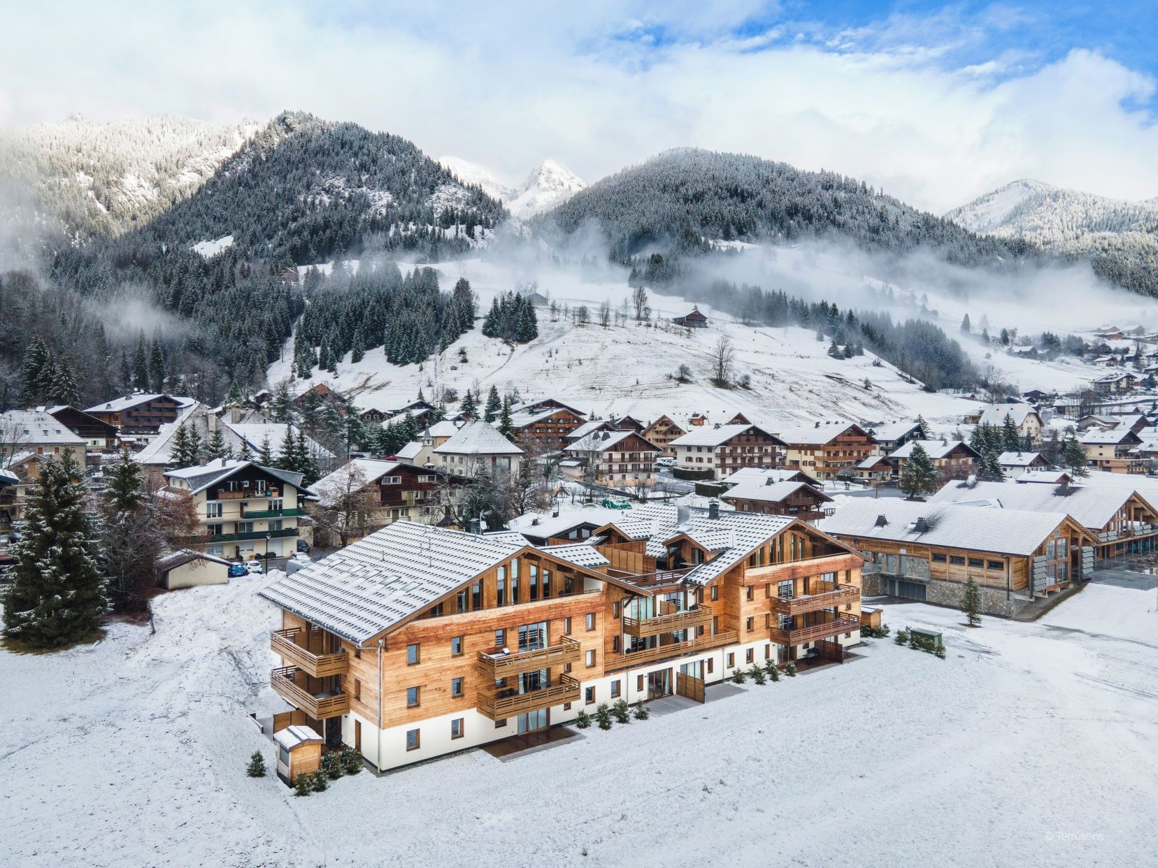 Ein verschneites Bergdorf mit Holzhäusern, darunter ein großes Chalet mit mehreren Balkonen und einer Skipiste im Hintergrund.