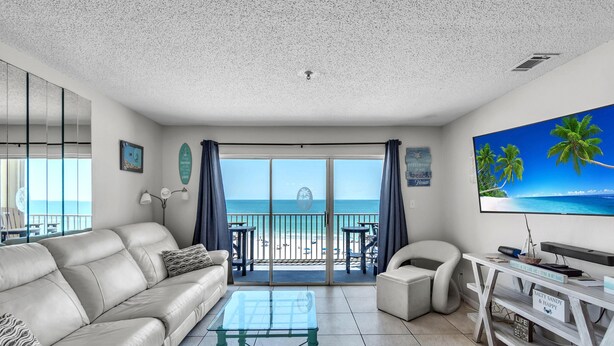A living room with a sofa, coffee table, and TV. There is a balcony with a view of the beach and palm trees. - Sea Breeze 503 (Madeira Beach)